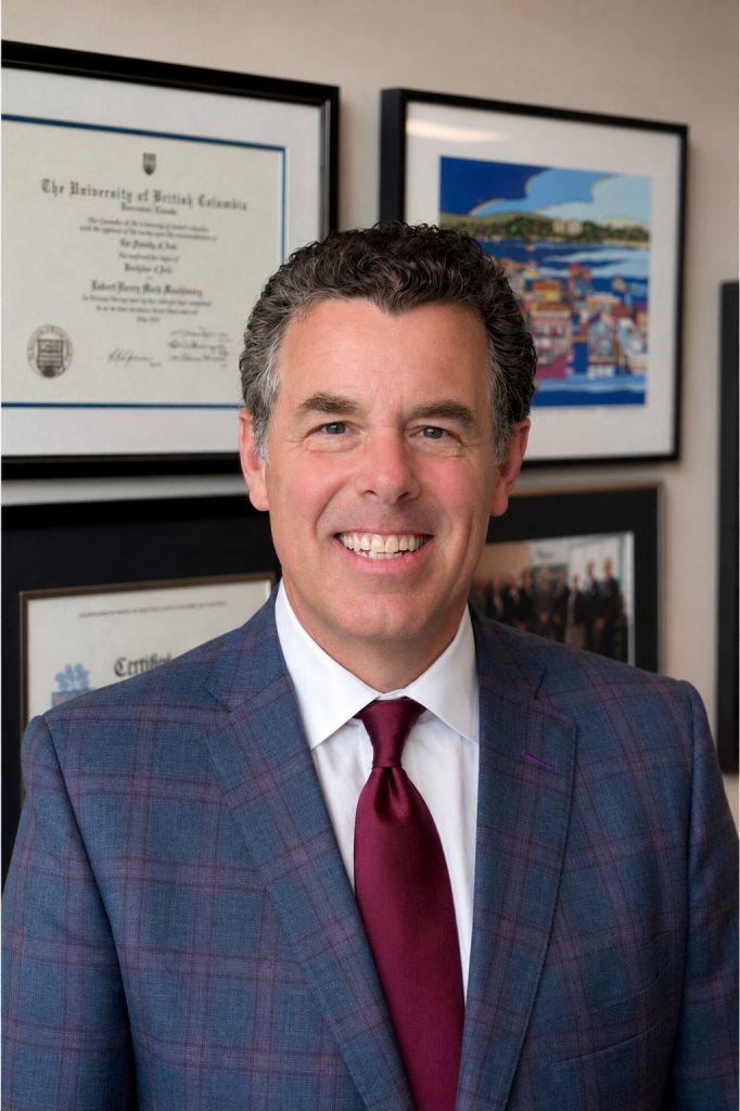 Professional head-and-shoulders portrait of a person wearing a blue plaid suit jacket, white shirt, and burgundy tie, standing in an office with framed certificates and artwork visible on the wall behind.
